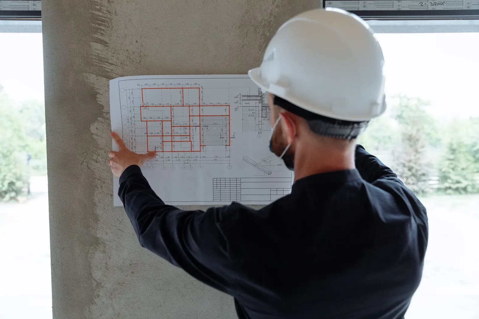 Planer mit Bauhelm prüft einen Grundrissplan an einer Betonstütze